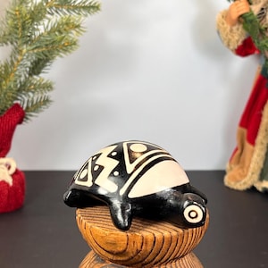 Puede incluir: Una figura de tortuga de cerámica en blanco y negro con diseños geométricos. La tortuga descansa sobre una base de madera. Un pequeño árbol de Navidad en el fondo.