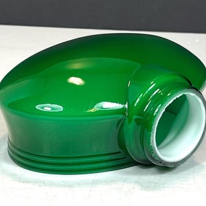 Könnte beinhalten: Ein glänzender grüner Glaslampenschirm mit abgerundeter Oberseite und zylindrischer Öffnung. Der Lampenschirm hat eine weiße Innenseite. Das Design weist eine glatte Oberfläche und eine geschwungene Form auf, passend für eine Vintage- oder Retro-Ästhetik. Der Lampenschirm ist ein leuchtendes Grün.