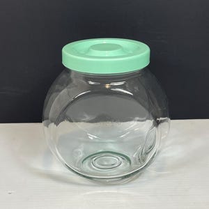 May include: A clear glass jar with a round shape and a light green lid. The jar is empty and sits on a white surface against a black background. The lid is a solid, opaque color.