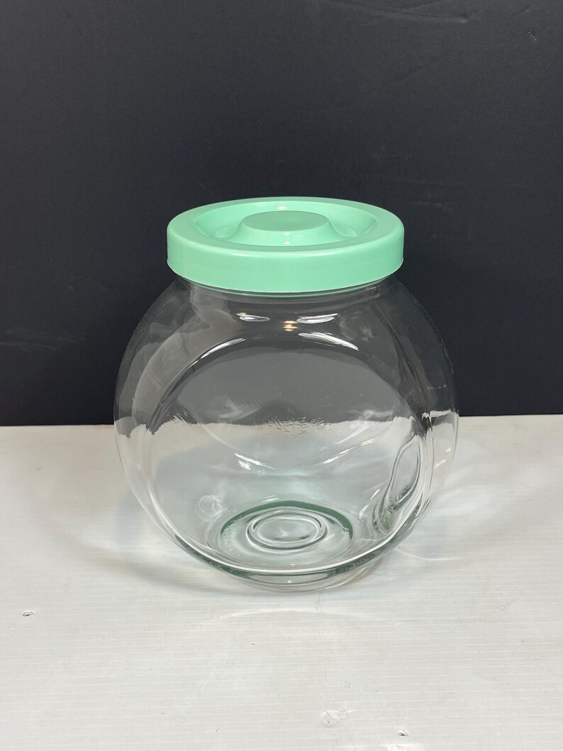 May include: A clear glass jar with a round shape and a light green lid. The jar is empty and sits on a white surface against a black background. The lid is a solid, opaque color.