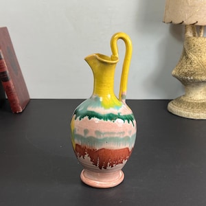 Könnte beinhalten: Ein bunter Keramikkrug mit gelbem Griff und Ausguss. Der Körper des Kruges weist horizontale Bänder in Rosa, Grün und Braun auf. Der Krug hat einen abgerundeten Boden und einen gebogenen Griff. Der Krug ist ein dekorativer Gegenstand.
