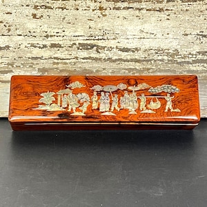 Puede incluir: Una caja rectangular de madera marrón rojizo con un acabado brillante. La tapa está adornada con una escena incrustada detallada con figuras, nubes y follaje en tonos blancos y crema. La caja se encuentra sobre un fondo claro y envejecido.