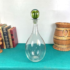 Peut inclure: Une carafe en verre transparent en forme de goutte d'eau, surmontée d'un bouchon vert en forme de sphère. Le bouchon présente un motif texturé en forme de bulles. La carafe est posée sur une surface turquoise, avec des livres et un panier tressé en arrière-plan.