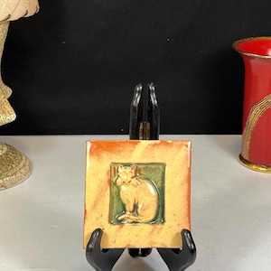 Puede incluir: Azulejo cerámico cuadrado con relieve de gato. El gato es de color naranja claro, sobre un fondo verde y enmarcado por un borde naranja y amarillo. El azulejo se exhibe sobre un soporte negro.
