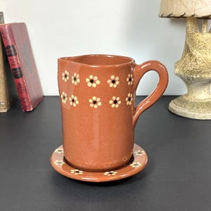 May include: A terracotta-coloured ceramic jug with a matching saucer. The jug features a handle and is decorated with small, white and yellow daisy-like flower designs. The saucer also has the same floral pattern around its edge.