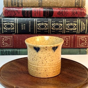 May include: A speckled beige ceramic pot with two blue heart designs near the rim. The pot sits on a wooden tray, with a stack of vintage books in the background. The books have red, black, and green covers.