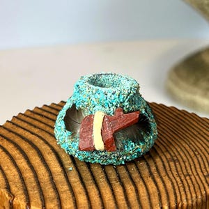 Puede incluir: Pequeño jarrón decorativo turquesa cubierto de pequeñas piedras trituradas. El jarrón tiene un diseño rectangular con detalles en rojo y amarillo y está adornado con plumas. Se encuentra sobre una base redonda de madera con una superficie texturizada y acanalada.