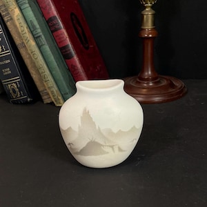 May include: A small white ceramic vase with a grey mountain scene painted on the front. The vase has a rounded body and a slightly flared lip.