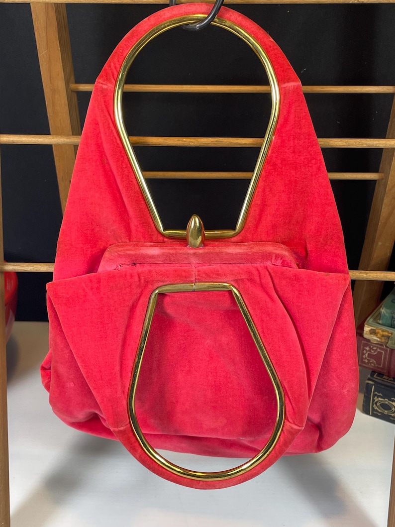 May include: A bright red velvet handbag with a gold metal top handle. The handle is shaped like an oval and has a pointed top.