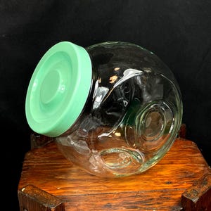 May include: A clear glass jar with a round body and a light green lid. The jar has a decorative design on the side and is sitting on a wooden surface. The jar is empty.