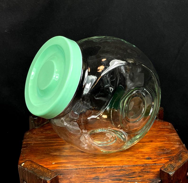May include: A clear glass jar with a round body and a light green lid. The jar has a decorative design on the side and is sitting on a wooden surface. The jar is empty.