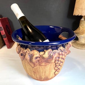 May include: A ceramic wine cooler with a dark blue rim and handles, holding a wine bottle. The beige body is decorated with grape and leaf relief carvings. A wine bottle is inside the cooler.