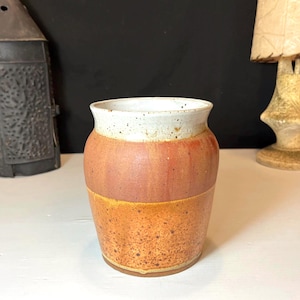 May include: A handmade ceramic vase with a speckled white rim, a reddish-brown middle section, and a textured orange-brown base. The vase has a rounded shape and is likely used for decorative purposes.