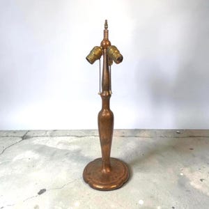 May include: A metallic bronze-toned table lamp base featuring a slender, contoured central column and a circular, scalloped base. The top assembly includes two light bulb sockets, each equipped with a vertical pull chain for independent operation.