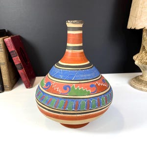 Op de afbeelding: Een handbeschilderde keramische vaas met een bolvormige body en een smalle hals. De vaas heeft een levendig ontwerp met banden in oranje, blauw, zwart en groen. De decoratieve vaas is een uniek stuk woondecoratie.