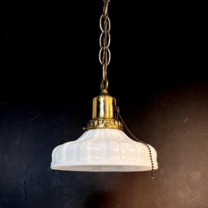 May include: A pendant light fixture featuring a white, fluted glass shade attached to a brass-toned metal socket and cap. The fixture is suspended by a matching brass-toned metal chain and includes a small beaded pull chain for operation.