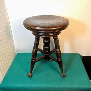 May include: A dark brown, antique wooden stool with a round seat and four legs. The legs are intricately carved and end in small, claw-like feet. The stool is likely vintage and adds a touch of classic charm.