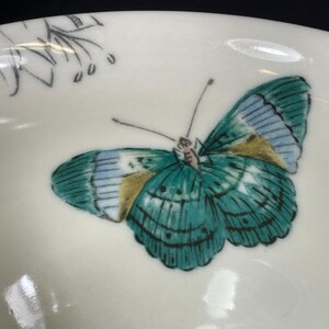 Op de afbeelding: Een witte keramische kom met een groen en blauw vlinderontwerp. De vlinder heeft gouden accenten op zijn vleugels.