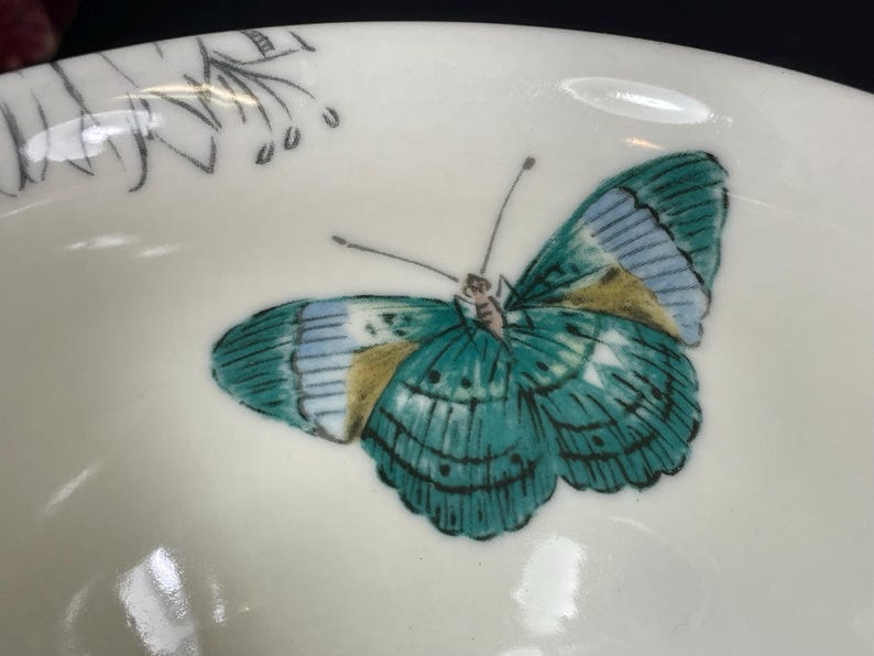 Op de afbeelding: Een witte keramische kom met een groen en blauw vlinderontwerp. De vlinder heeft gouden accenten op zijn vleugels.
