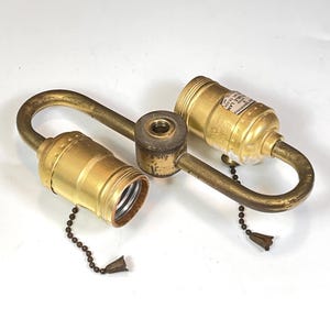 May include: A vintage brass lamp fixture with two bulb sockets. The fixture has a curved design with a central mounting point. Each socket has a pull chain with a decorative tassel. The brass has a slightly aged appearance.