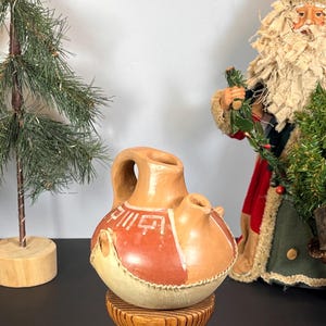 Puede incluir: Una jarra de cerámica de color canela con asa y dos picos. La jarra tiene un diseño rojo y canela con líneas blancas y una banda cosida similar al cuero alrededor de la base. Se encuentra sobre un soporte de madera, con un pequeño árbol de Navidad y una figura de Papá Noel en el fondo.