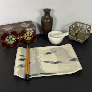 May include: A set of Asian-inspired home decor items including a wooden box with intricate floral designs, a white ceramic mortar and pestle, a small metal cage, a brown ceramic vase, and a white ceramic plate with a painted design and two wooden chopsticks.