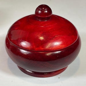 May include: A round, wooden container with a lid, finished in a deep red stain. The container has a bulbous shape with a small, round knob on top of the lid. The base is a slightly darker shade of red, and the wood grain is visible.