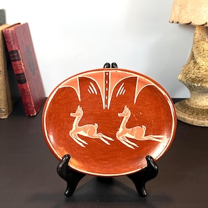 May include: A terracotta-colored ceramic plate with a decorative design. The plate features two stylized deer in white and peach, set against a background of geometric shapes. The plate is displayed on a black stand.