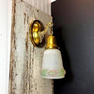 Może przedstawiać: Zabytkowy mosiężny kinkiet z owalną płytą tylną i ozdobnym ramieniem. Oprawa oświetleniowa posiada klosz z matowego szkła z zielono-różowym kwiatowym wzorem. Zamontowany na zwietrzałej drewnianej powierzchni.