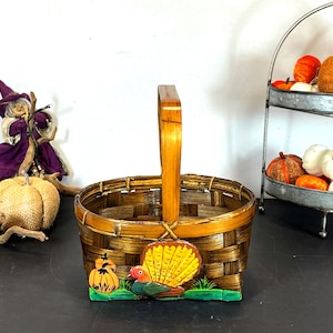 May include: A woven brown basket with a wooden handle. The basket features a decorative panel with a turkey, pumpkins, and green grass. A witch figurine and pumpkins are in the background, along with a tiered metal tray filled with decorative pumpkins.