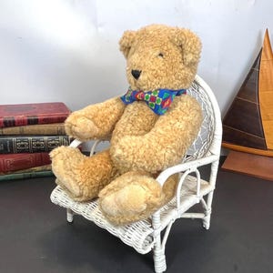 May include: A light brown teddy bear wearing a patterned bow tie sits in a white wicker chair. The chair is on a dark surface. A stack of old books is to the left of the bear. The bear has a soft, plush appearance.