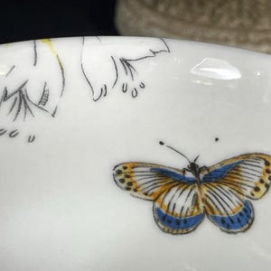 Op de afbeelding: Een witte keramische kom met een bloemenpatroon en een blauwe en gele vlinder.