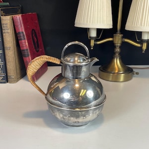 Może przedstawiać: Srebrny czajnik z wiklinowym uchwytem. Czajnik ma okrągły korpus i dzióbek. Stoi na białym tle.