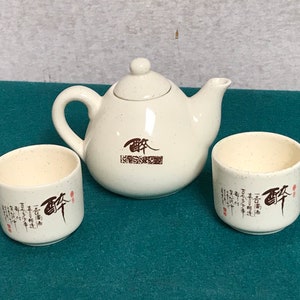 Puede incluir: Una tetera de cerámica blanca con una inscripción marrón y dos tazas de té a juego. La tetera tiene un cuerpo redondo y un pequeño pico. Las tazas de té son pequeñas y redondas. La inscripción en la tetera y las tazas de té está en japonés.