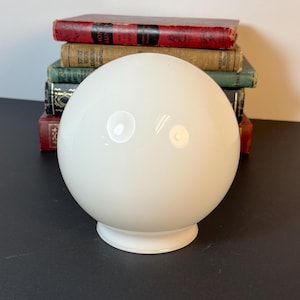 Könnte beinhalten: Ein glatter, kugelförmiger Lampenschirm aus weißem Glas mit einer kreisförmigen Öffnung an der Basis. Der Schirm steht vor einem Stapel aus fünf alten Hardcover-Büchern mit verschiedenfarbigen Buchrücken in Rot, Beige, Grün und Schwarz mit Golddruck.