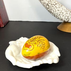 May include: A decorative, hand-painted egg with a yellow and brown base, adorned with floral and heart designs in red and gold. The egg rests in a white, open-hand shaped dish. The background includes books and a mid-century modern lamp.