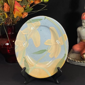 May include: A round ceramic plate with a light blue background and yellow floral design. The plate is supported by a black stand. The flowers have green stems and leaves. A vase of fall foliage is in the background.