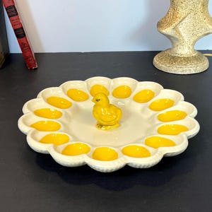 May include: A white ceramic egg tray with a scalloped edge, featuring yellow egg-shaped indentations and a small yellow chick figurine in the center. The tray is designed to hold eggs and is a decorative kitchen item.