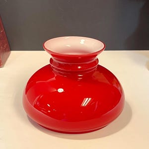 May include: A bright red glass vase with a white interior. The vase features a rounded base, a cylindrical neck, and a flared rim. The glossy finish reflects light, highlighting its smooth surface. A decorative piece for home decor.