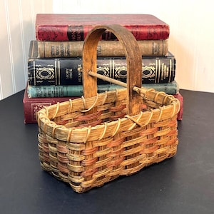 May include: A rectangular woven basket featuring a tall, arched wooden handle and a woven base made of light brown and reddish-brown strips. The basket has a sturdy, open-top design with a cross-woven pattern and a smooth, curved wooden handle.