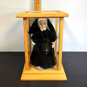 Puede incluir: Muñeca de porcelana de una monja con hábito y velo negros, expuesta dentro de una vitrina de madera y cristal. La muñeca tiene una toca blanca y un collar con una cruz dorada. La vitrina es rectangular.