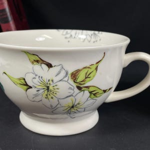 Op de afbeelding: Een witte keramische theekop met een bloemenontwerp met witte bloemen met gele centra en groene bladeren. De kop heeft een afgeronde vorm en een handvat.