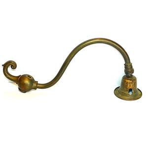 May include: An antique brass-toned wall sconce arm with a curved design. The arm features a decorative floral-like detail on one end and a bell-shaped light socket on the other. The metal has a weathered appearance.