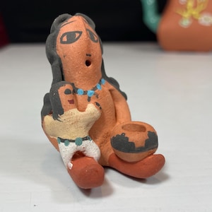 Puede incluir: Figurita de cerámica pintada a mano de una mujer nativa americana sosteniendo a un niño. La mujer lleva un collar turquesa y el niño lleva un vestido blanco con un cinturón turquesa. La figurita está sentada sobre una superficie blanca.