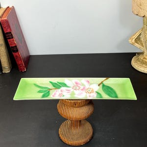 Op de afbeelding: Een rechthoekige, lichtgroene keramische schaal met een bloemenontwerp. De schaal heeft witte en roze bloemen met groene bladeren en bruine stelen. De schaal wordt op een houten standaard gepresenteerd.