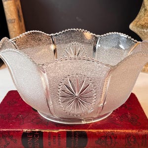 Peut inclure: Un bol en verre transparent doté d'une surface texturée granuleuse et d'un bord festonné. L'extérieur du bol est orné de motifs d'étoiles pressées répétitifs à l'intérieur de cadres ovales, créant un contraste visuel net avec la texture givrée.