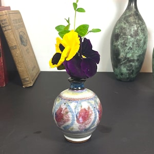 May include: A small, round ceramic vase with a floral design, holding a yellow and purple pansy. The vase has a blue rim and a white base. Two other vases and several books are in the background.