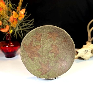May include: A green ceramic plate with a brown leaf design. The plate is on a white surface and is being held up by a metal stand.
