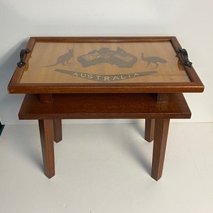 May include: A wooden tray table with a glass top featuring a graphic of the Australian coat of arms. The tray table has two black leather handles and sits on a wooden stand with four legs.