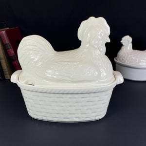 Peut inclure: Plat à couvercle en céramique blanche en forme de coq avec une base en osier. Un plat plus petit en forme de poule est également visible. Les plats sont sur une surface sombre avec des livres en arrière-plan.